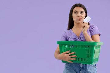 Poster - Thoughtful young woman with credit card and shopping basket on lilac background