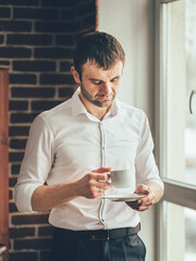 Wall Mural - A man in a white shirt is holding a cup of coffee. He looks sad and is looking out the window