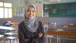 © Radiant Art - Asian school teacher wearing hijab standing in a classroom chairs tables and board behind her
