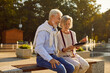© Studio Romantic - Portrait of happy smiling senior couple sitting outdoors on bench in city park using digital tablet for social media, searching information, watching funny movie or having online video call.