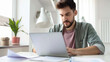 © VK Studio - A bearded man sits at a desk, intently working on a laptop, surrounded by papers and modern office decor with natural light streaming in.