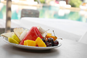 Wall Mural - Plate with fresh fruits on table near sun lounger, space for text. Luxury resort with outdoor swimming pool