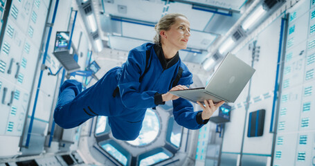 Female Astronaut Using Laptop Computer in Space Inside a Spacecraft. Working Online, Checking and Updating Flight Information or Writing Report. Spacewoman Floating in Zero Gravity Inside Spaceship