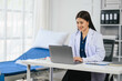 © PaeGAG - Asian doctor in a white lab coat is sitting at a desk with a laptop. She is smiling and she is happy
