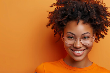 Wall Mural - A woman with curly hair smiles brightly while wearing round glasses and an orange shirt, standing against a bright orange backdrop