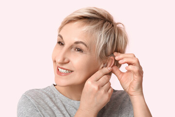 Poster - Mature woman putting hearing aid on her ear on pink background