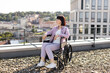 © sofiko14 - Young woman in wheelchair enjoying outdoor rooftop view on a sunny day. She is dressed casually in a light purple outfit and exudes a sense of calm and contemplation while appreciating the cityscape.