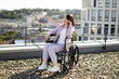 © sofiko14 - Woman in wheelchair enjoying a sunny day on an urban rooftop. She appears relaxed and contemplative while taking in the cityscape. The backdrop of buildings and greenery adds to the serene mood.