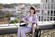 © sofiko14 - Smiling woman sitting in wheelchair on rooftop terrace with city view in background. Dressed in casual clothing, looks comfortable and content. Concept of accessibility, independence, and lifestyle.