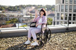 © sofiko14 - Confident woman sitting in wheelchair on rooftop terrace, smiling and making peace sign on sunny day. Modern urban cityscape in background. Embracing positivity and independence.