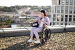 © sofiko14 - Woman in wheelchair holds coffee and smartphone while enjoying a scenic view of city. The setting is outdoors on a rooftop with a backdrop of urban buildings.
