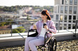 © sofiko14 - Woman in wheelchair enjoys outdoor rooftop view while holding cup of coffee and smartphone. Relaxed and happy, she connects with nature and cityscape.