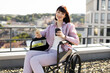 © sofiko14 - Smiling woman in wheelchair enjoying coffee and using smartphone on a rooftop with cityscape in background. Bright day, relaxed moment, urban setting.