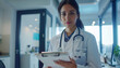 © Oleksiy - Portrait of young latin american female doctor wearing white coat standing in clinic office, filling out medical form at clipboard