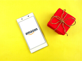 Closeup amazon com app on white phone screen & red gift box, gold ribbon bow. Amazon logo of online electronic shop website, red gift box on yellow background top view