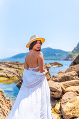 Wall Mural - A woman wearing a white dress and a straw hat stands on a rocky beach