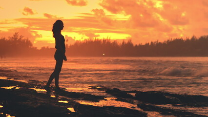 Woman stands alone on a sandy beach sets in background, casting a warm glow over horizon. The sky vibrant oranges, pinks, and purples, creating a stunning silhouette of the woman against colorful sky