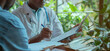 © Muhammad - doctor sitting his desk holding empty clipboard writing one hand while talking patient front of him closeup shot realistic photography style using bright color natural light There green plant