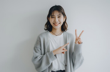 Wall Mural - Japanese woman her early mid twenty smiling giving peace sign one hand wearing business casual attire of light gray cardigan over white shirt black jean against clean white background She standing