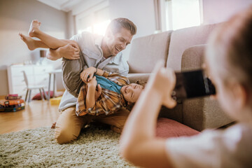 Wall Mural - Father playing with son while being photographed by daughter at home