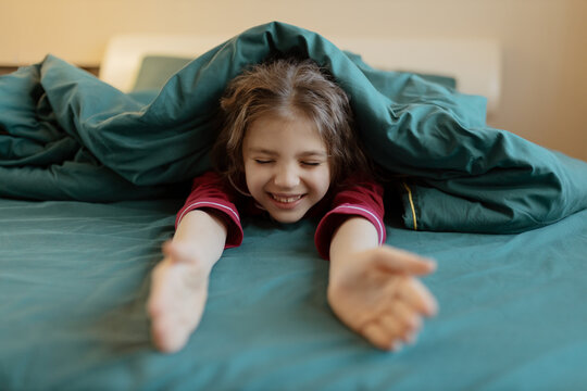 Happy 7 year old girl in red pyjamas in green blanket arms outstretched on bed at home