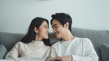 Wall Mural - young asian couple wearing light  clothing, looking contented and happy. They are looking into each others eyes reclining on a grey sofa. White col background with uncluttered elements. Overall image 