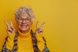 Happy senior woman in glasses, wearing yellow, making peace sign with both hands, bright background

