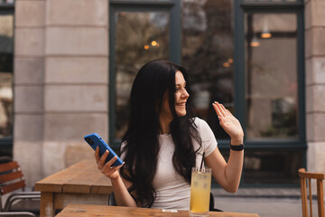 Beautiful young woman looking at side hold mobile phone sitting in cafe and wave her hand, sign hi, hello. Brunette girl in white t-shirt sitting on the chair in restaurant and called waiter