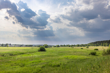 A wide shot of a green meadow with a few trees scattered throughout. The sky is dominated by large, dark clouds with patches of sunlight breaking through.