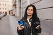 © zvkate - Beautiful smiling woman walking on street with coffee takeaway to go, chatting on mobile phone. Girl talking on smartphone on her way to meeting spot, sending message, wear black leather jacket.