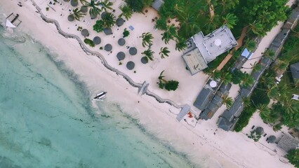 Wall Mural - Aerial view of green palm trees, waves, umbrellas on the sandy beach of Indian Ocean at sunset. Summer in Zanzibar island. Tropical landscape with green palms, white sand, clear sea. Top drone view