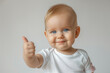 © mankjon - Cheerful baby with light skin and round blue eyes giving a thumbs-up, wearing a white shirt, isolated on a plain background, capturing the essence of happiness and positivity in early childhood