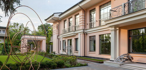 Sticker - Modern luxury suburban house with pale peach walls and large bay windows, complemented by a garden with artistic metal sculptures.