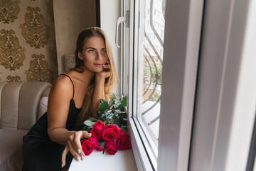 Wall Mural - A woman in a black dress is sitting in front of a window with a bouquet of red roses.