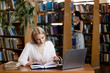 © Kateryna Muzhevska - a girl sits at a table in a library, reading a book and taking notes. There is also a laptop and several closed books on the table.