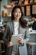 © pham - A smiling businesswoman in a gray blazer stands in a modern cafe, holding a tablet in her hands