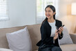 © amnaj - A cheerful woman sitting on a couch, smiling and holding her smartphone, dressed in a casual black and white outfit, enjoying her time at home.