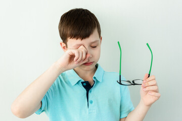 Small school guy rubs his eyes standing on gray background. Dry eyes, pain. Bad eyesight, fatigue