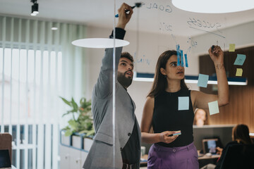 Wall Mural - Two professional coworkers, a man and a woman, actively engage in brainstorming, using sticky notes on a glass wall to organize ideas and strategies in a well-lit contemporary office.