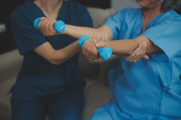 Old woman training with physiotherapist using dumbbells at home. Therapist assisting senior woman with exercises in nursing home. Elderly patient using dumbbells with outstretched arms.