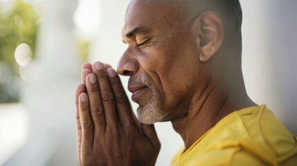 Man with closed eyes hands clasped in prayer wearing a yellow shirt standing against a blurred background with a hint of greenery and a white wall.