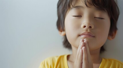 Wall Mural - Young child with eyes closed hands clasped together in prayer wearing a yellow shirt against a soft-focus background.