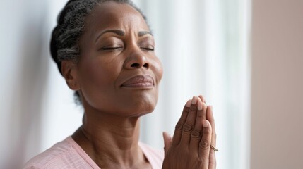 A woman with closed eyes hands clasped in prayer standing in front of a window.