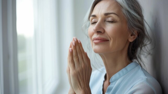 Woman with closed eyes hands clasped in prayer standing in front of a window wearing a light blue top with a serene expression.