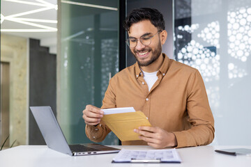 Wall Mural - Happy indian man sitting at desk in modern office and smilingly opening and reading received letter