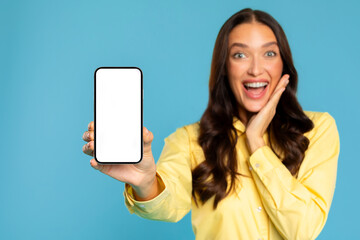 Amazing offer. Excited surprised European lady showing cellphone with white empty screen and exclaiming with joy, standing over blue studio background, mockup