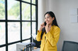 © PaeGAG - Young Professional Woman in Yellow Blazer Talking on Smartphone in Modern Office with Large Windows and Natural Light