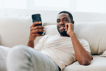 Happy African American man sitting on a black sofa in his modern apartment, typing a message on his mobile phone He is dressed in a white tshirt and exudes confidence as he connects with friends and