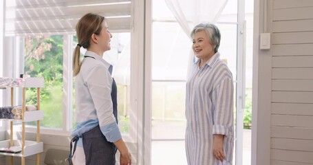 Poster - Asian girl, small business spa shop owner, standing to welcome customers at door. Girl in shirt and denim apron smiling, greeting senior customer in striped dress, emphasizing hospitality and warmth.