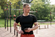 © arthurhidden - A fit, athletic man takes a break after a workout, holding his smartphone and wearing wireless earbuds. He stands confidently at an outdoor fitness park, showcasing active lifestyle.